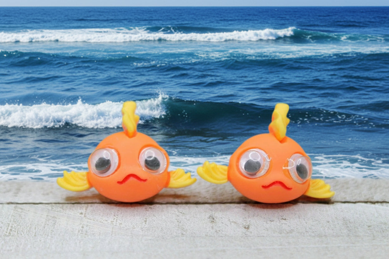 Two orange fish-shaped earrings with yellow fins on a wooden surface with a green blurred background.