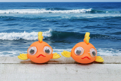 Two orange fish-shaped earrings with yellow fins on a wooden surface with a green blurred background.